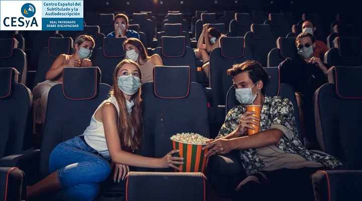 En un cine con gente con mascarilla, una pareja en primera fila comparten un bol de palomitas con una butaca vacía en medio. 