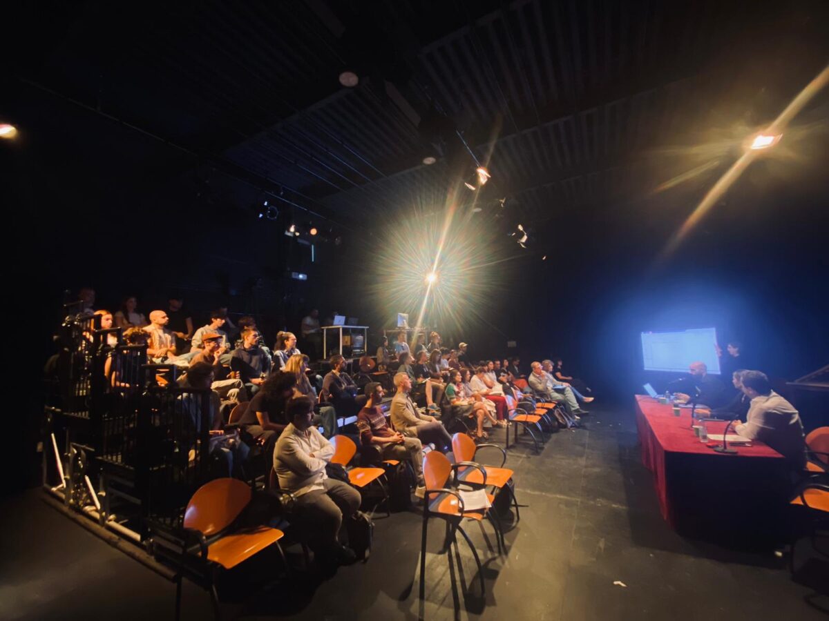 Panorámica del auditorio con el público en grada frente a la mesa de bienvenida. 